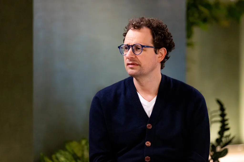 A man in glasses and a dark cardigan sits looking to the side in a softly lit office