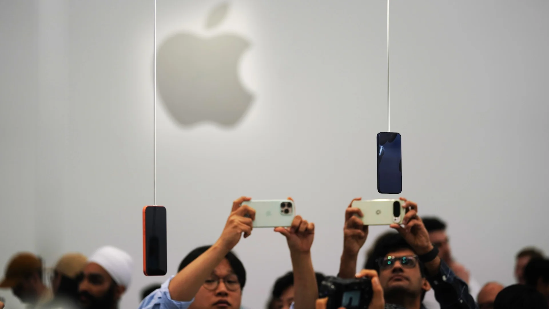 People raise phones to photograph iPhones suspended on wires beneath a large Apple logo at a launch event