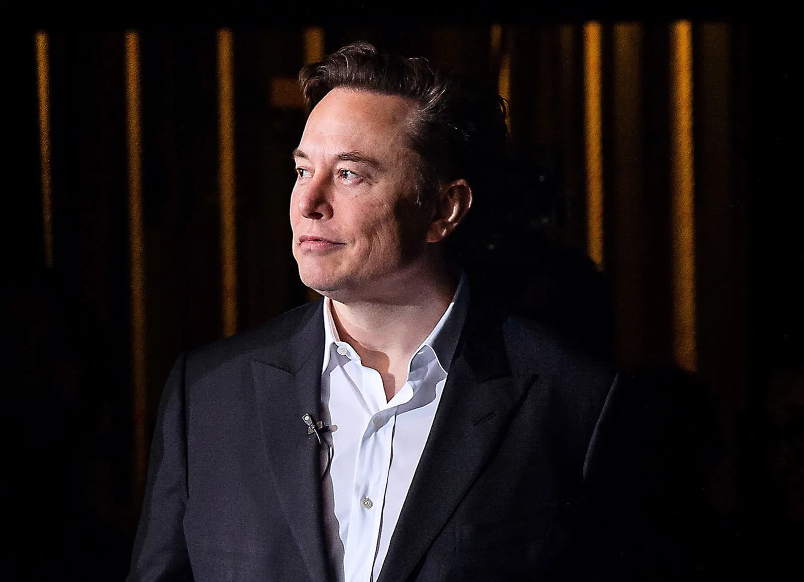 Elon Musk in a dark suit looking to the side against a shadowed background