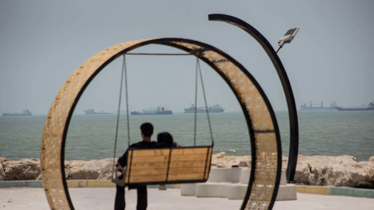 Cargo ships anchored offshore near an Iranian port seen past a seafront swing