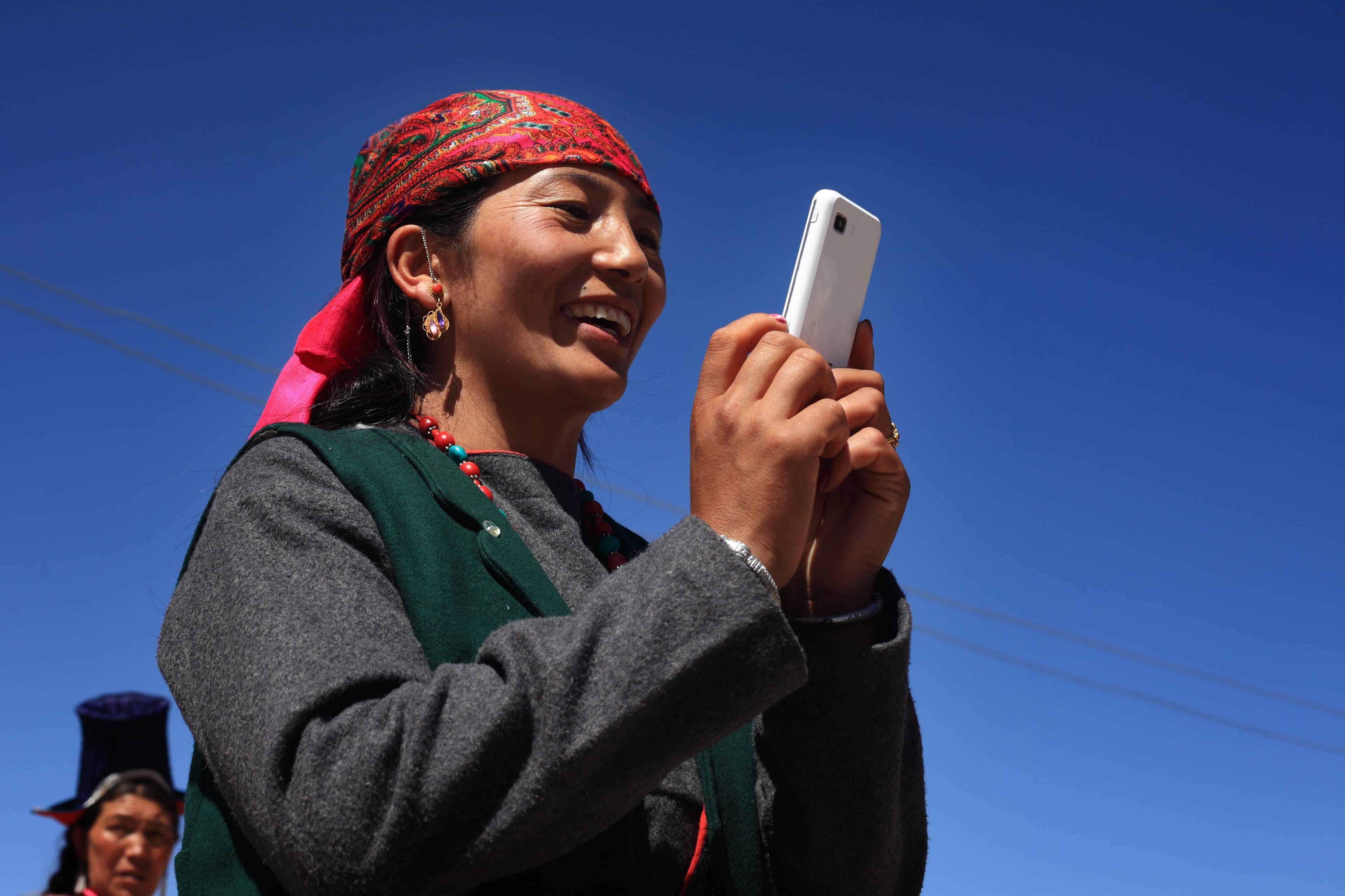 A woman in a bright headscarf smiles as she looks at a smartphone under a clear blue sky