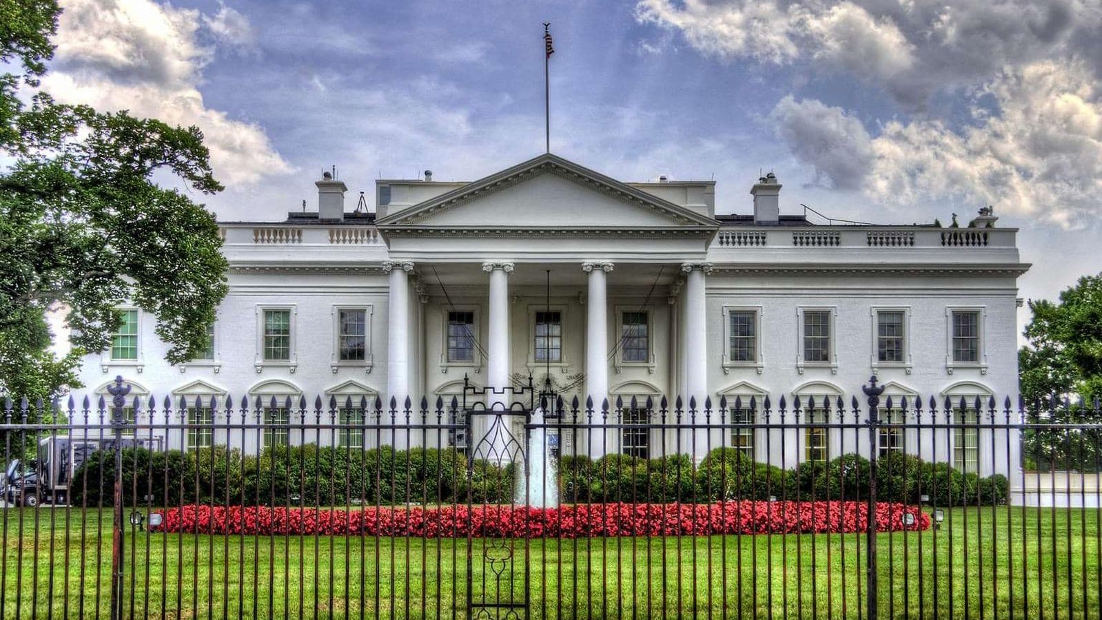 The White House seen from the north behind its black iron fence, with red flowers along the green lawn