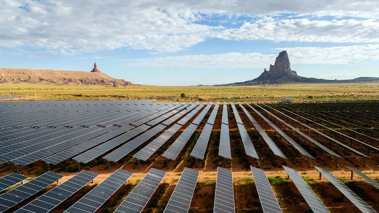 Aerial view of a large solar panel farm spread across red desert land beneath rock formations