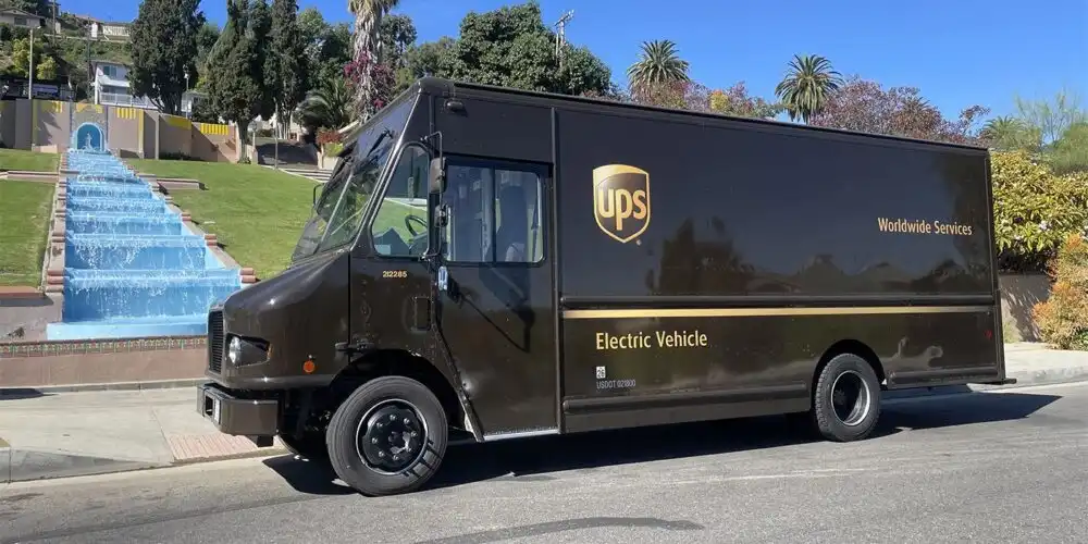A brown UPS electric delivery van parked on a sunny street beside a stepped water fountain