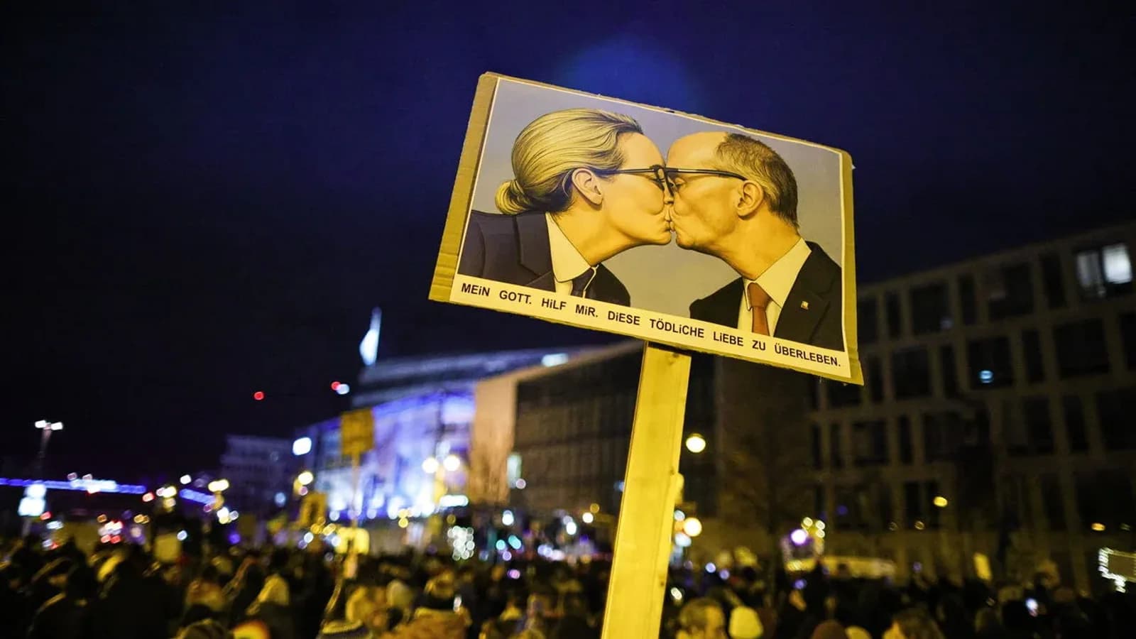 A protest placard parodying the socialist fraternal kiss, depicting the AfD and CDU leaders kissing, held above a night-time crowd in Germany