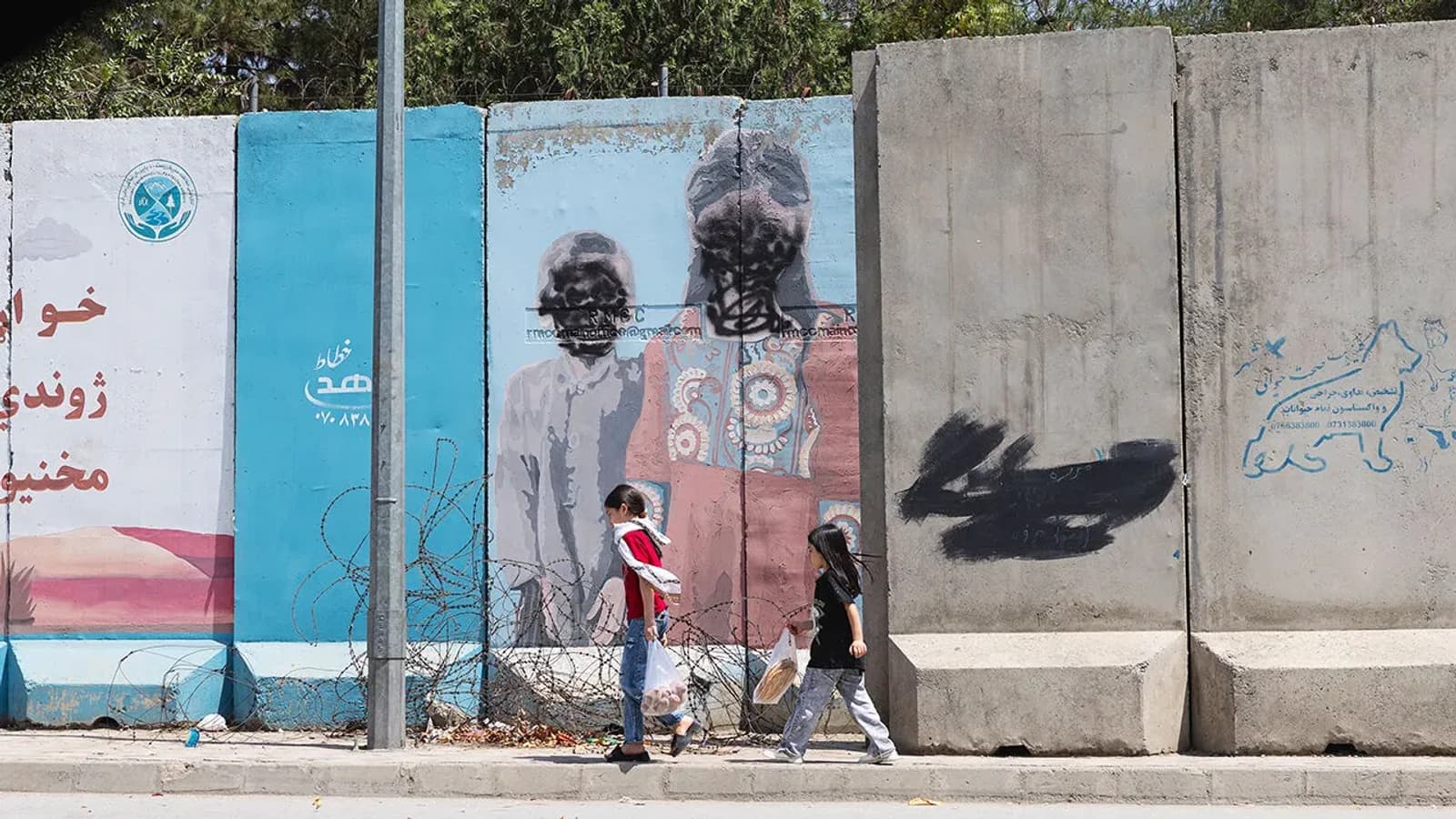 Two girls carrying shopping bags walk past a defaced mural and concrete blast walls on a Kabul street