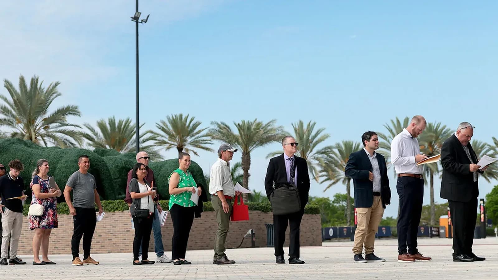 A long line of job seekers holding papers waits outdoors at a hiring event, with palm trees behind them