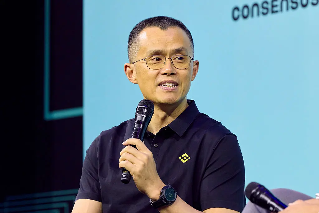 A man in glasses holds a microphone and speaks on stage in front of a blue backdrop reading Consensus