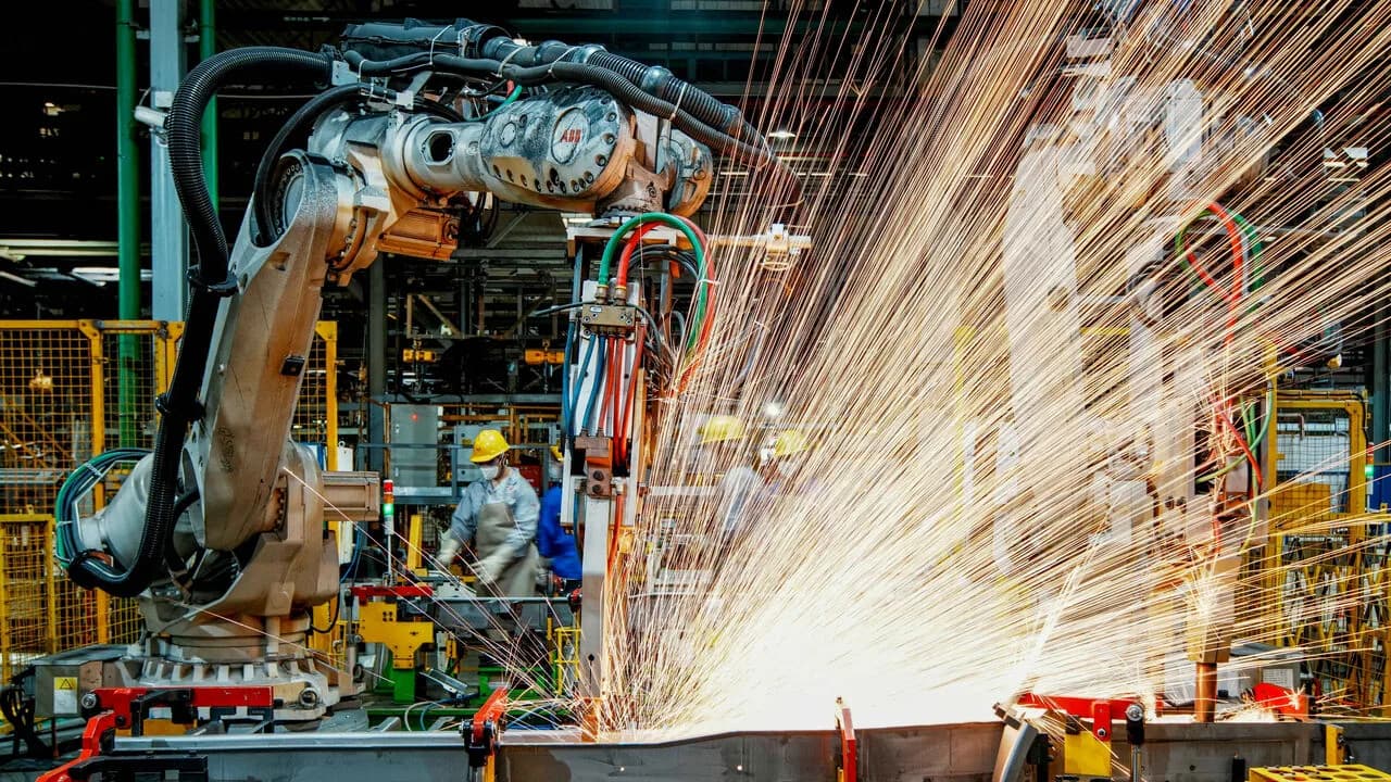 An industrial welding robot throws a shower of sparks over an assembly line as workers look on inside a Chinese factory