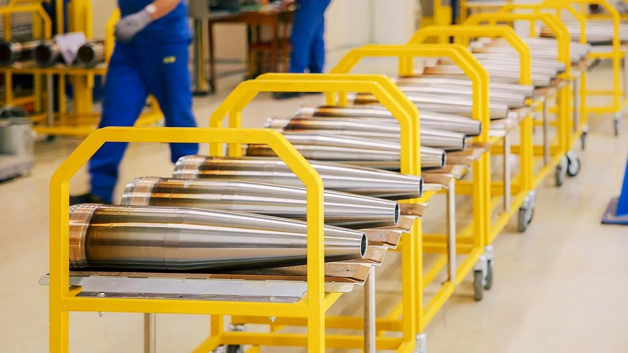 Rows of freshly machined large-calibre artillery shells on yellow trolleys inside a munitions factory, with workers in blue overalls
