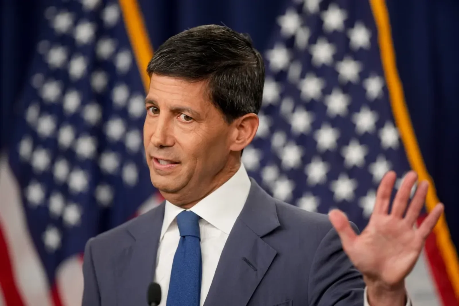 Kevin Warsh gestures with a raised hand while speaking at a podium in front of American flags