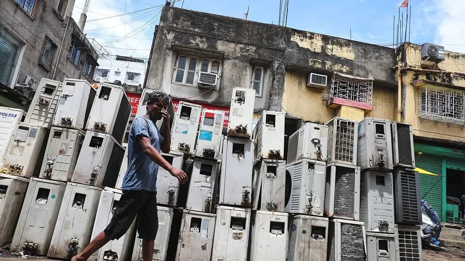 A man walks past a towering stack of discarded air conditioning units piled against old buildings in a hot Indian city