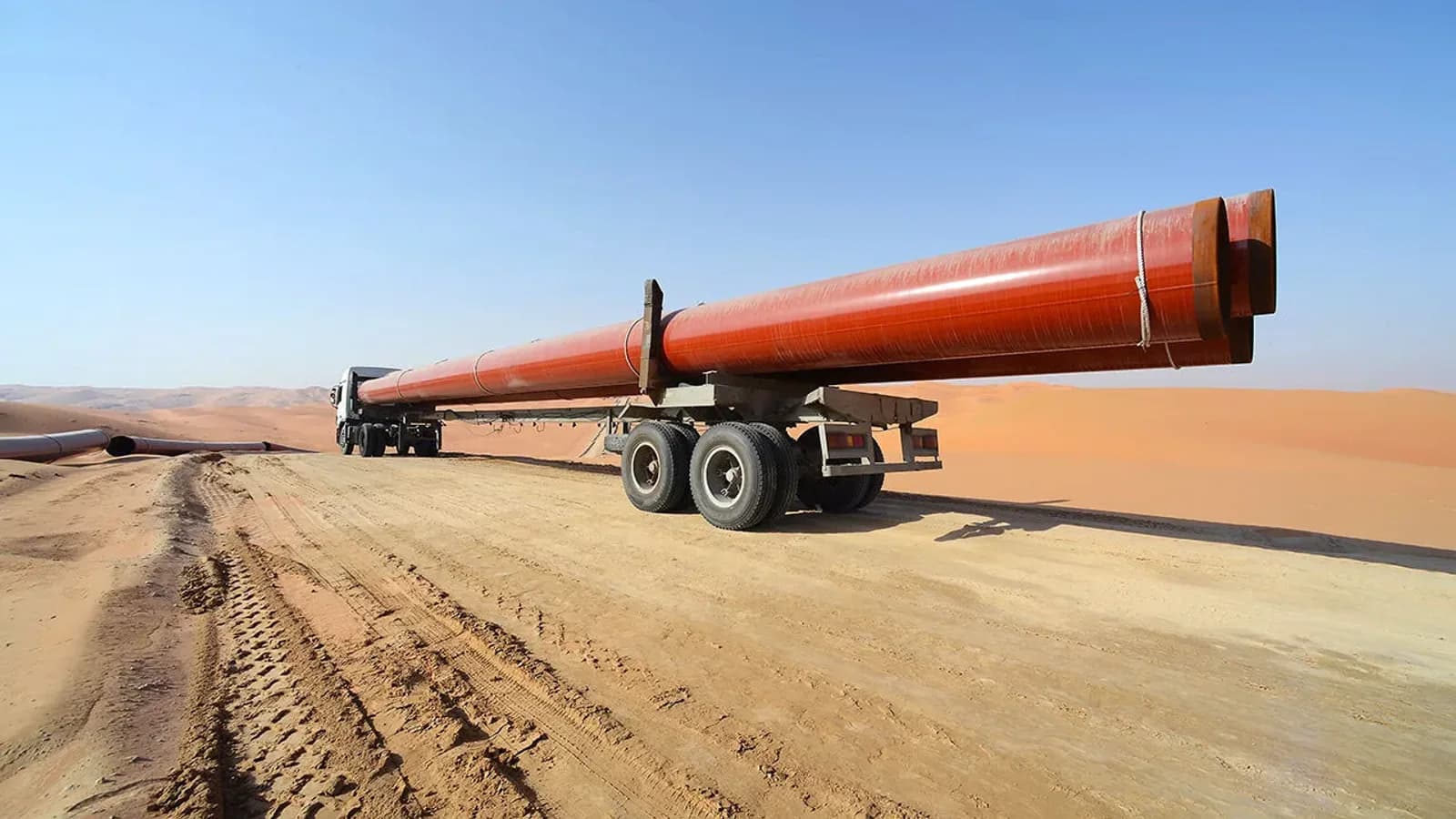 A truck hauls a long section of red oil pipeline across desert sand under a clear sky