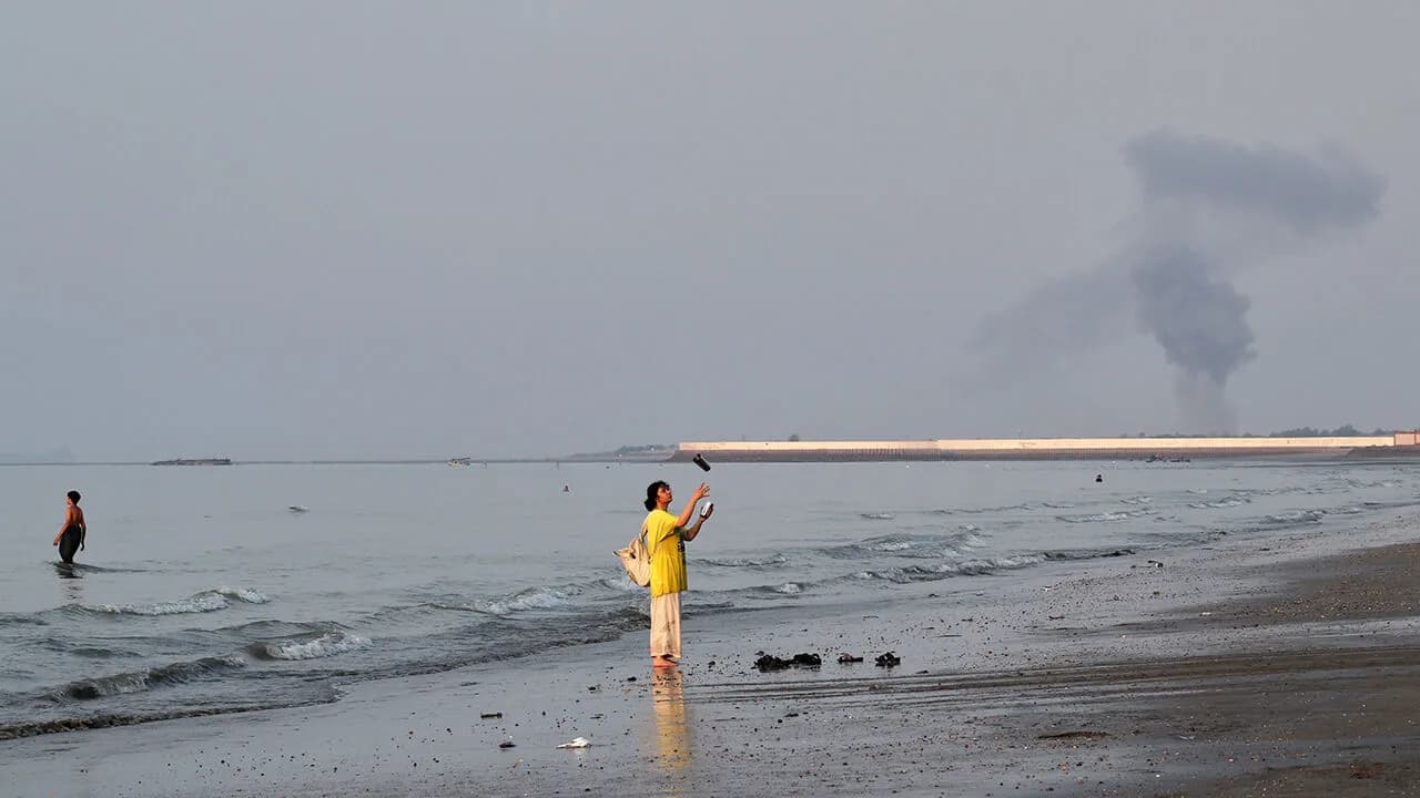 A man stands on a Gulf beach as a plume of dark smoke rises on the horizon during the Strait of Hormuz crisis
