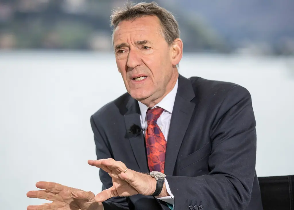 Jim O'Neill gestures with both hands while speaking during a television interview, wearing a dark suit and red tie