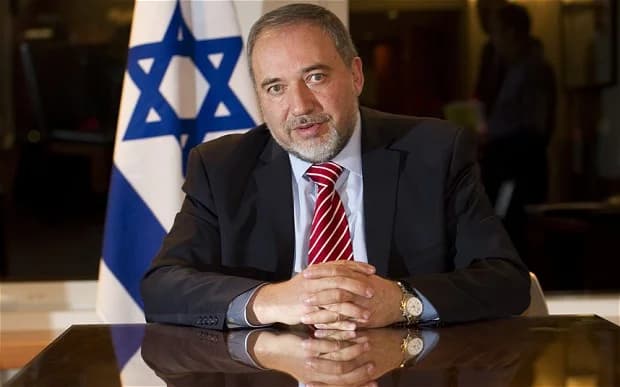 Avigdor Lieberman sits at a desk with hands clasped, an Israeli flag visible behind him