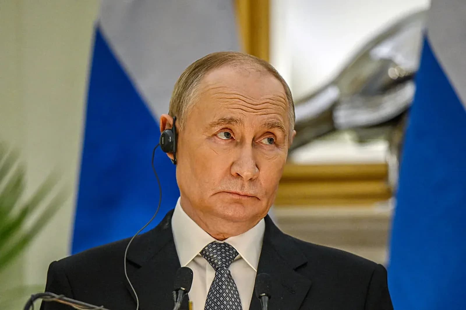 Vladimir Putin wearing a translation earpiece at a press conference, with a blurred Russian flag behind him