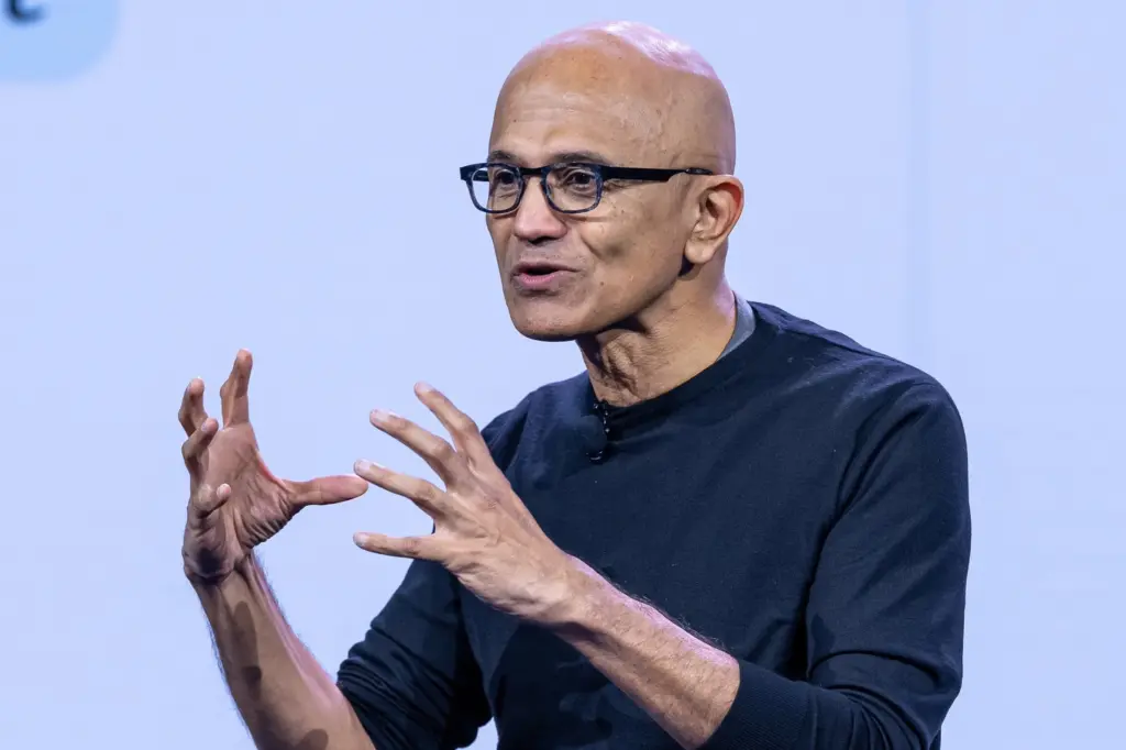 Satya Nadella gestures with both hands while speaking on stage, wearing glasses and a dark sweater