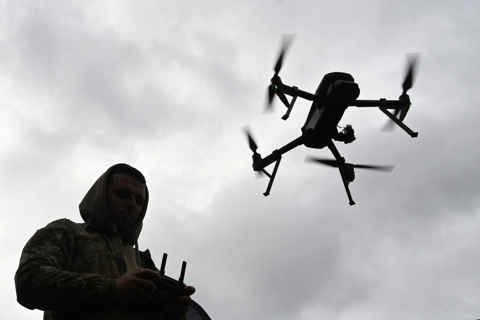 Silhouette of a soldier in camouflage operating a reconnaissance drone that hovers against a grey overcast sky