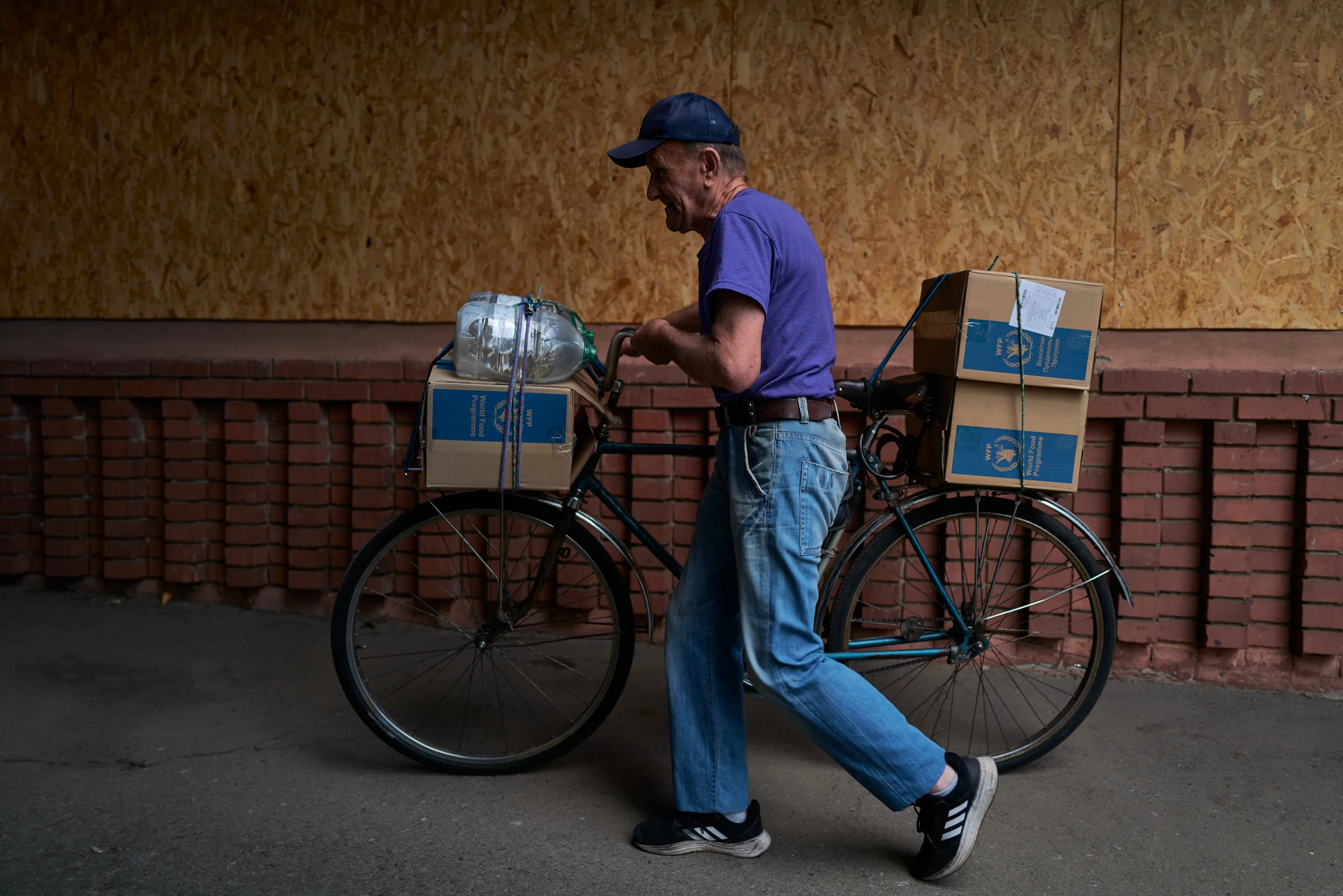 An elderly man wheels a bicycle loaded with World Food Programme aid boxes and bottled water past a boarded-up building in a Ukrainian frontline town