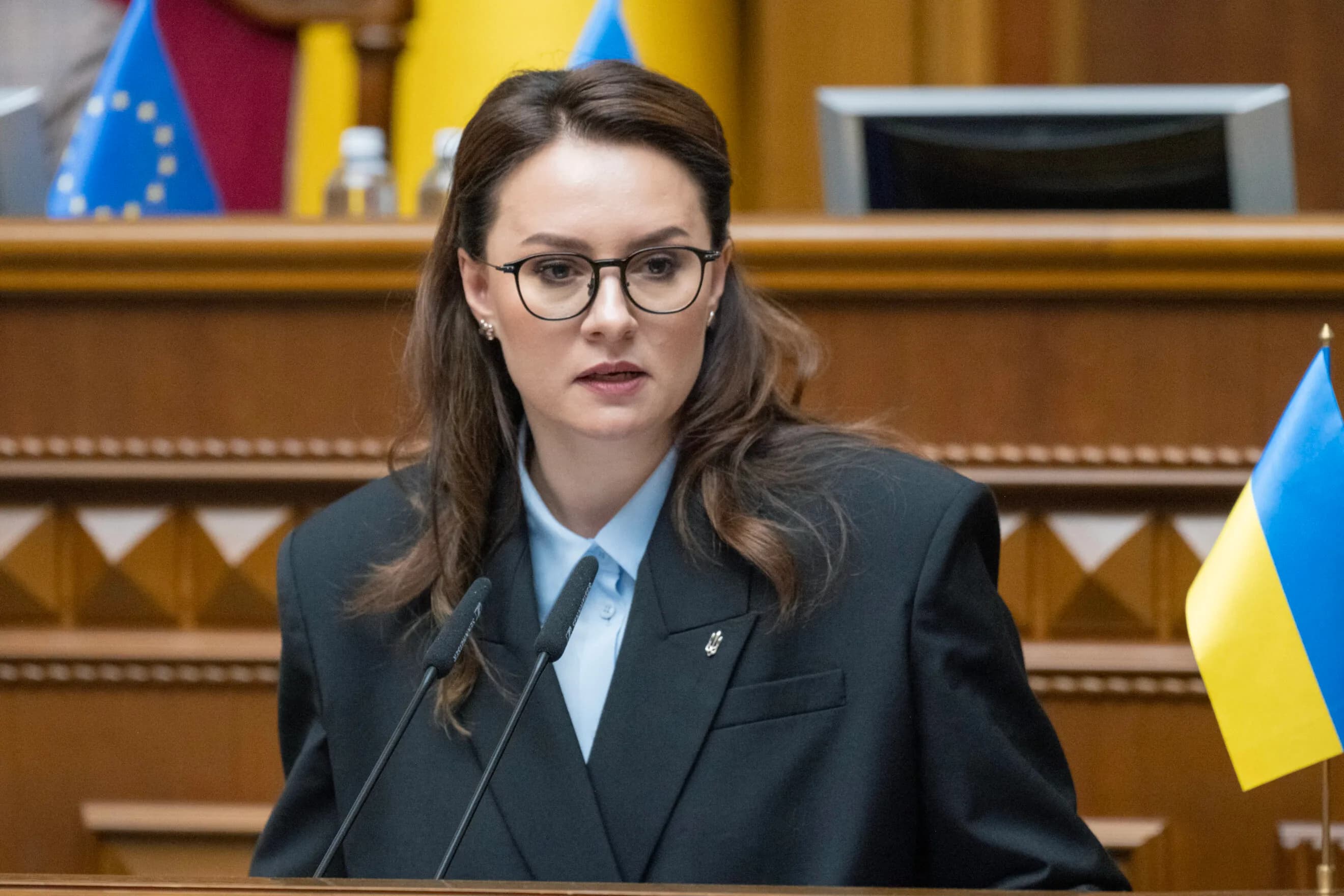 Ukrainian Prime Minister Yulia Svyrydenko speaks at the podium of the Verkhovna Rada, with Ukrainian and EU flags behind her