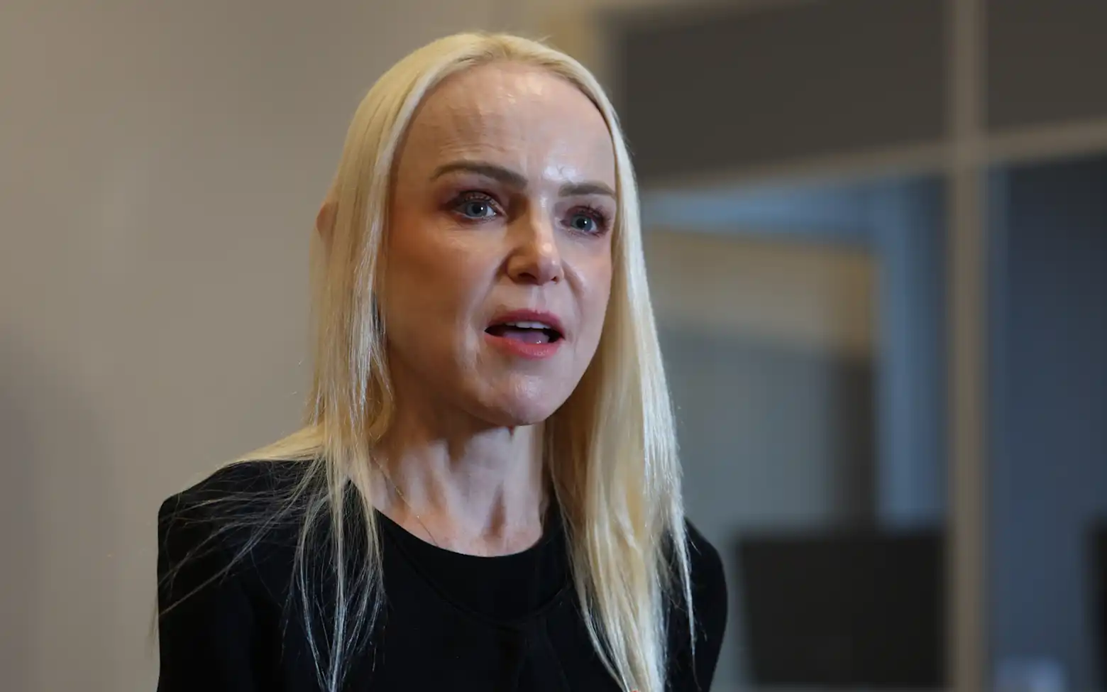 A blonde woman in a black top speaks during an interview, with a blurred office interior in the background